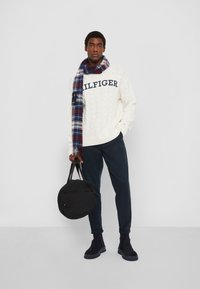 Mann in weißem Kabelstrickpullover mit "HILFIGER"-Schriftzug, kariertem Schal, marineblauen Hosen und schwarzen Stiefeln, der eine schwarze runde Tasche vor einem schlichten Hintergrund hält.