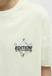 Camiseta de algodón amarillo claro con un diseño gráfico que incluye el texto "EDICIÓN" y "TODOS LOS DERECHOS RESERVADOS". Textura suave, escote redondo.