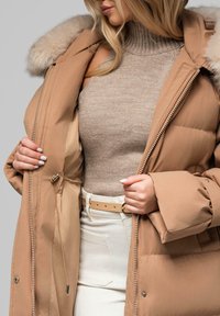 Piumino in tessuto marrone chiaro con cappuccio foderato in pelliccia, chiusura con zip, tasche laterali e cordino regolabile. Indossato sopra un maglione di maglia beige.