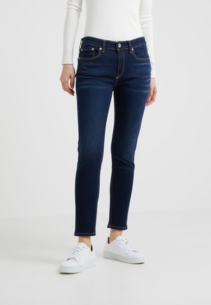 Frau trägt dunkelblaue Skinny-Jeans, weißes Langarmshirt und weiße Turnschuhe, steht vor einem schlichten weißen Hintergrund.