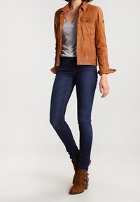 Chaqueta de ante color marrón con cierre de botones y bolsillos en el pecho, camiseta gris, jeans ajustados azul oscuro y botines de tacón marrón con detalles de hebilla.