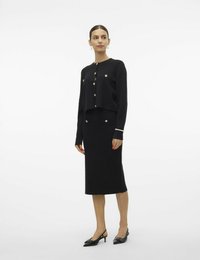 Cardigan corto nero con bottoni dorati abbinato a una gonna a tubino nera. Gli accessori includono un bracciale di perle e décolleté neri con cinturino.