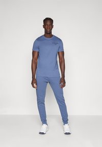 Modré bavlněné tričko a odpovídající tepláky, slim fit. Tričko má malé černé logo; tepláky mají žebrované manžety a boční kapsy.