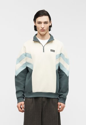 Mladý muž oblečený v krémovo-tyrkysové mikině s čtvrtzipem a vzorem cikcak, s tmavě hnědými kalhotami, stojící před jednobarevným světlým pozadím.