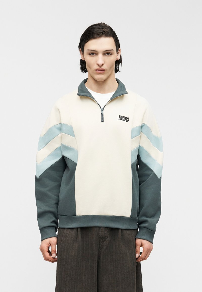 Νεαρός άνδρας που φοράει εκρού και πετρόλ φούτερ με φερμουάρ στο ένα τέταρτο και σχέδια σε σχήμα βέλους, καθώς και σκούρο καφέ παντελόνι, στέκεται μπροστά σε απλό φωτεινό φόντο.