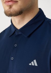 Marinblå polo skjorta med en strukturerad krage och knäppningar; har en grå logotyp på vänster sida nära bröstet. Slät tygkvalitet.