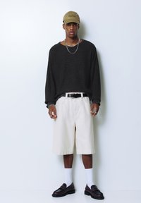 Donkere oversized trui, crèmekleurige korte broek tot de knie, olijfgroene pet en bruine loafers met zwarte accenten. Accessoires zijn een ketting en ringen.
