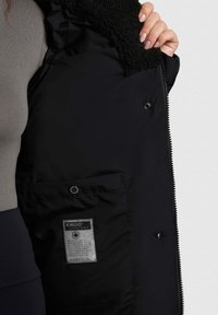 Cappotto isolato nero con colletto morbido e peloso, chiusura con zip e tasca laterale con etichetta contenente le istruzioni per la cura.