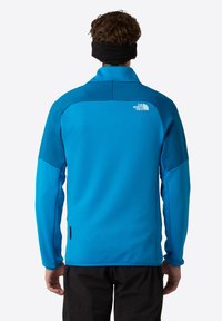 Giacca softshell blu con maniche lunghe, colletto alto e pannelli spalla a contrasto. Presenta un logo sulla parte superiore della schiena. Texture liscia.