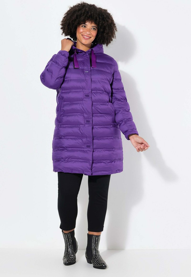 Lila Pufferjacke mit Kapuze, mit horizontalen Steppungen, Knopfverschluss und dekorativen Bändern. Getragen mit schwarzen Leggings und verzierten Stiefeletten.