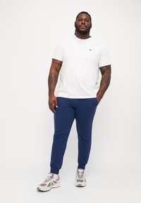 Hvit t-skjorte med korte ermer og en liten rød logo, kombinert med blå joggebukser og grå sneakers med mørke detaljer.
