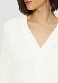 White blouse with a V-neckline and long sleeves, featuring smooth fabric and a relaxed fit. The design is minimalistic with no patterns.