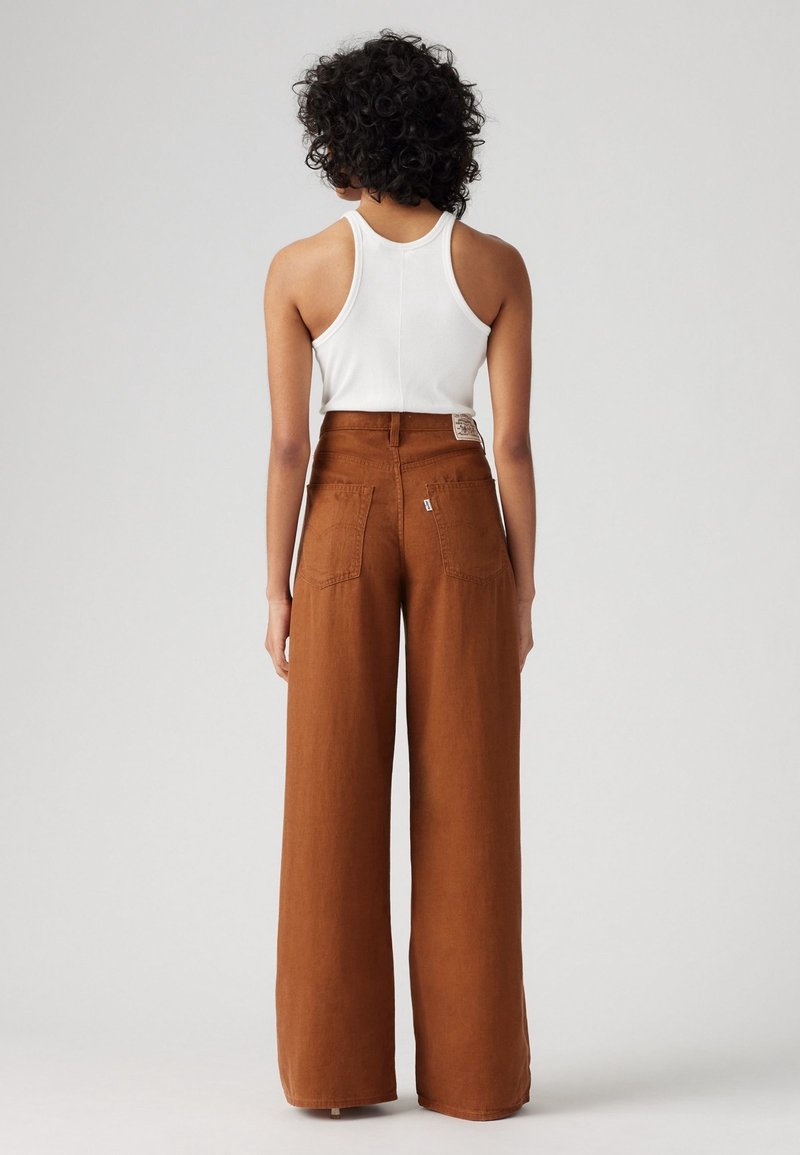 Personne aux cheveux courts et bouclés, de dos, portant un débardeur blanc et un pantalon marron à taille haute et jambes larges sur un fond uni.