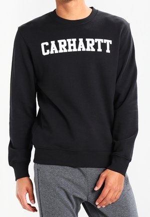 Homme portant un sweat-shirt noir Carhartt à col rond et un pantalon de jogging gris, debout devant un fond blanc.