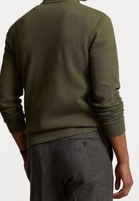 Olivgrüner Strickpullover mit langen Ärmeln, strukturiertem Stoff und lockerer Passform, kombiniert mit maßgeschneiderten grauen Hosen mit einer Gesäßtasche.