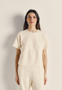 Korte mouw, crème kleurige top gemaakt van gestructureerde stof met een ronde halslijn en een losse pasvorm. De zoom is iets korter gesneden.
