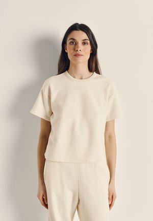 Krátky rukáv, smotanovo sfarbený top vyrobený z textúrovaného materiálu s okrúhlym výstrihom a voľným strihom. Lem je mierne skrátený.
