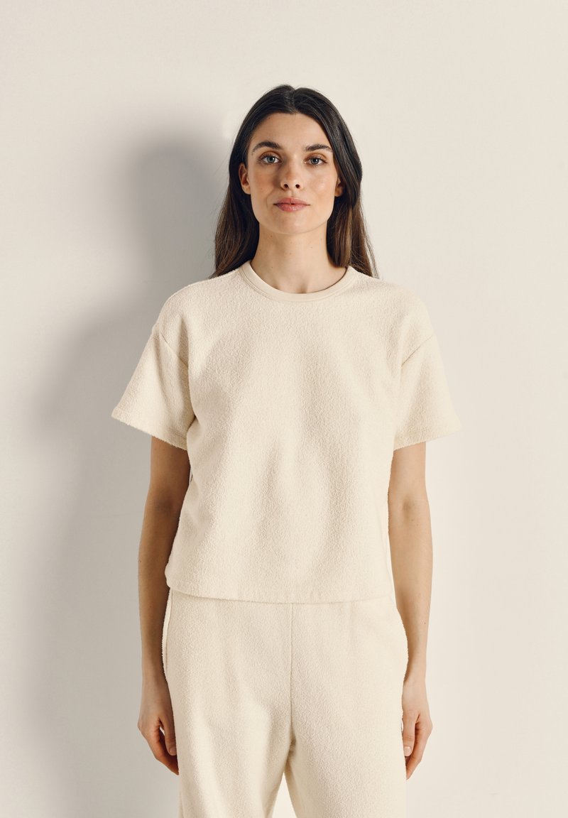 Korte mouw, crème kleurige top gemaakt van gestructureerde stof met een ronde halslijn en een losse pasvorm. De zoom is iets korter gesneden.