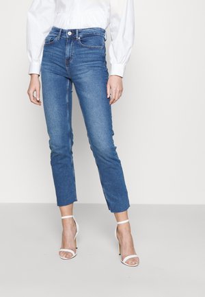 Kobieta w niebieskich obcisłych dżinsach z wysokim stanem, białej bluzce z długim rękawem i bufkami oraz białych sandałkach na wysokim obcasie z paskiem wokół kostki.