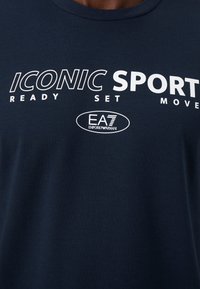 T-shirt de algodão azul-marinho com texto gráfico branco "ICONIC SPORT READY SET MOVE" e logo EA7 Emporio Armani. Textura lisa, decote redondo.