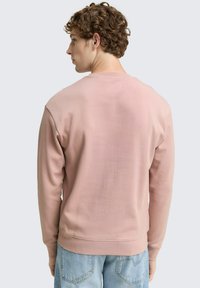 Felpa rosa chiaro con una texture liscia, scollo a girocollo e orlo a costine. Le maniche sono leggermente affusolate, indossata con jeans di denim blu chiaro.