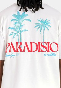 Witte katoenen t-shirt met rode tekst "PARADISIO" en blauwe palmboomgraphics. De tekst bevat "is callin..." en merkdetails daaronder.
