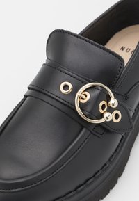 Mocasines de cuero negro con un hebilla circular decorativa en tono dorado, diseño en capas y detalles texturizados en la parte superior. Suela antideslizante.