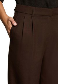 Pantalons plissés marron en tissu texturé, dotés de poches latérales et d'une taille ajustée, maintenus partiellement à la taille par une main.