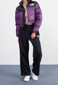 Woman wearing a purple and black cropped puffer jacket, beige crop top, black Nike sweatpants, and white sneakers standing on carpeted floor.