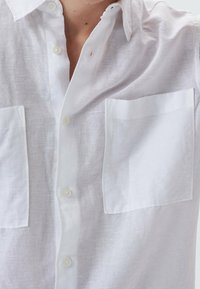 Primo piano di una persona che indossa una camicia di lino bianca con due tasche sul petto e chiusure a bottoni, in cui si vedono il colletto e la parte superiore del torso.