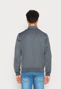 Chaqueta bomber gris con cuello y puños acanalados, material ligero y textura suave. Lleva encima unos pantalones vaqueros azules.