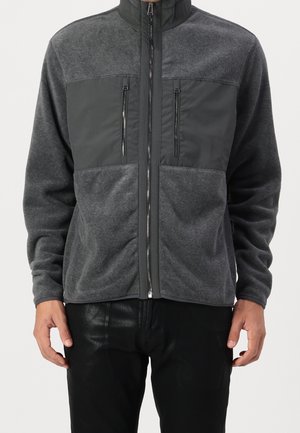 Mann trägt eine dunkelgraue Fleecejacke mit durchgehendem Reißverschluss, zwei vertikalen Reißverschlusstaschen auf der Brust und schwarze Hosen.