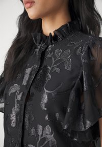 Femme portant un chemisier noir transparent avec des motifs floraux métalliques, col haut à volants et manches flottantes, montrée du cou à la poitrine.