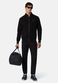 Giacca nera con zip e tasche laterali, abbinata a pantaloni neri e sneakers blu. Modello che tiene una borsa da palestra nera con un logo.