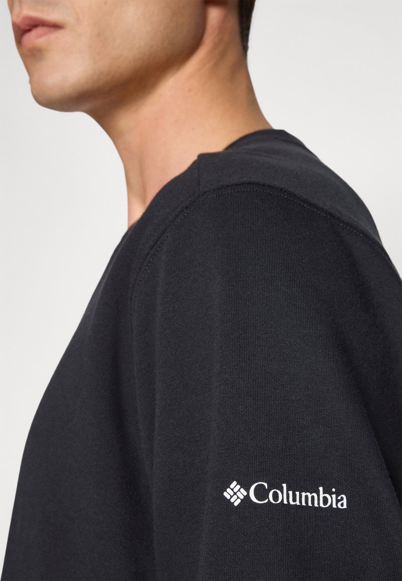 Sweat-shirt noir à col rond, arborant un logo Columbia blanc sur la manche gauche. Tissu doux avec une texture lisse et un design épuré.