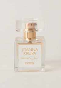 Szklana butelka perfum o kwadratowym kształcie, przezroczystym korpusie i złotym wieczku. Etykieta zawiera biały i złoty tekst.