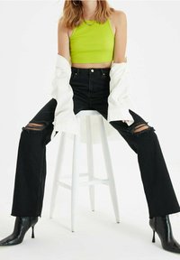 Donna seduta su uno sgabello bianco, indossa un top corto verde lime, jeans larghi neri strappati, una camicia bianca aperta, stivali neri con tacco e una collana d'oro.