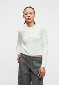 Biela pletená kardigán s knoflíkovým zapínaním a dlhými rukávmi, v kombinácii so sivými cargo nohavicami s viacerými vreckami a uvoľneným strihom.