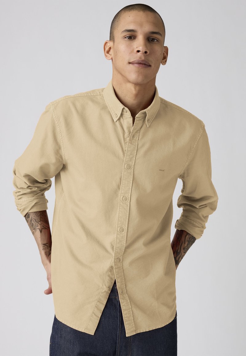 Junger Mann mit hochgekrempelten Ärmeln, der ein beiges Hemd mit Knöpfen und dunkle Jeans trägt, steht mit den Händen hinter dem Rücken vor einem einfarbigen Hintergrund.