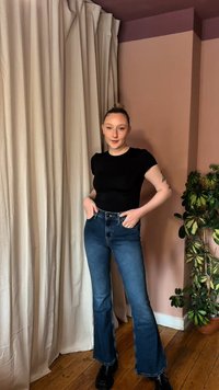 Νεαρή γυναίκα με μαλλιά σε κότσο, που φοράει μαύρο πουκάμισο, τζιν μπλε με ψηλή μέση και φαρδιά καμπάνα, και μαύρες μπότες, στέκεται δίπλα σε μπεζ κουρτίνες και ένα πράσινο φυτό.