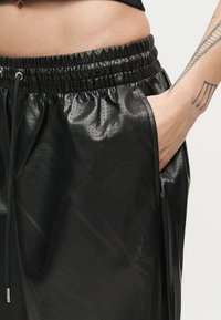 Prim-plan cu o persoană purtând pantaloni negri din piele ecologică perforată, cu talie elastică, mâna în buzunar și un tatuaj geometric pe antebraț.