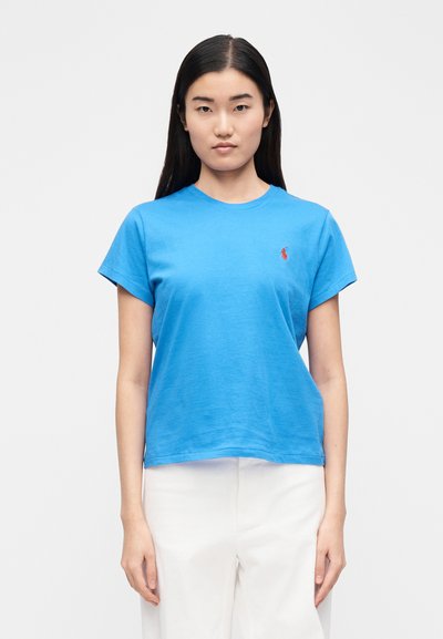 Mėlynas medvilninis marškinėlis su apvalia iškirpte, trumpomis rankovėmis ir mažu raudonu siuvinėtu logotipu kairėje krūtinės pusėje. Lygus audinys, laisvas prigludimas.