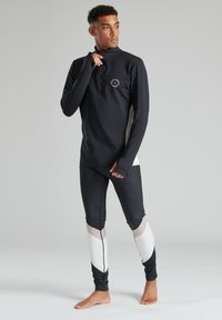 Schwarzes Langarmshirt mit hohem Kragen und Logo, kombiniert mit schwarzen Leggings mit weißen und beigen geometrischen Akzenten.