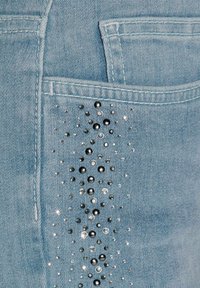 Helle blaue Jeans aus Denim mit einer Tasche, verziert mit verschiedenen metallischen Nieten und Strasssteinen, die ein strukturiertes Muster erzeugen.