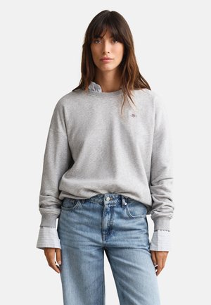 Žena s dlouhými hnědými vlasy má na sobě světle šedou mikinu přes bílou košili s límečkem, která je zastrčená do vysokých modrých džínů, stojí před bílým pozadím.