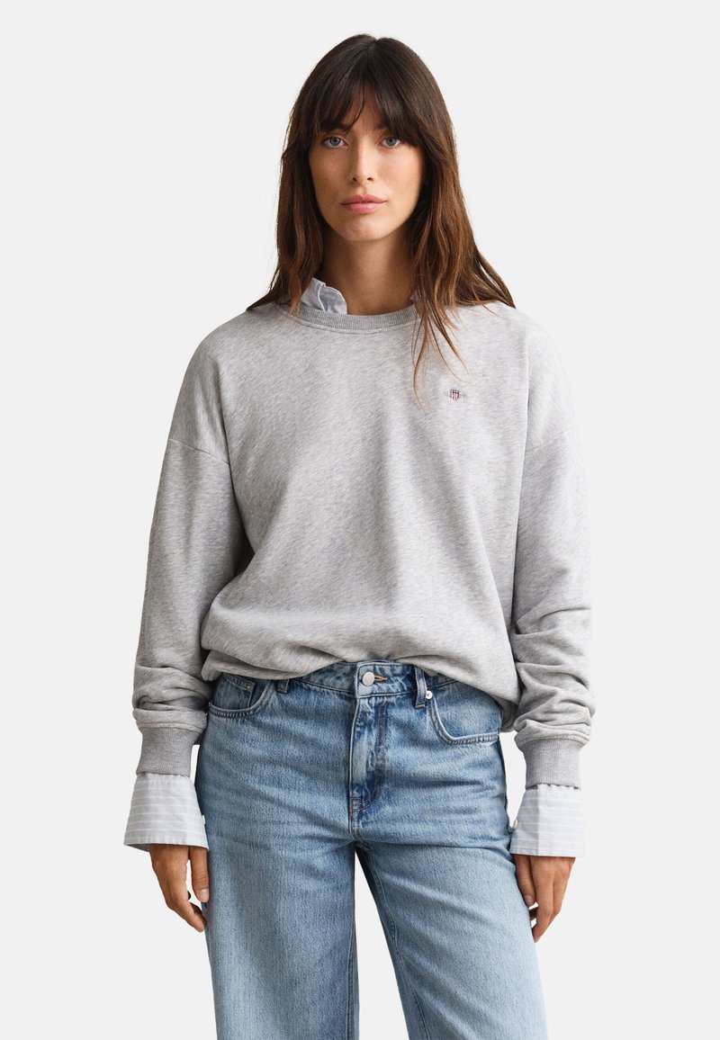 Femme aux longs cheveux bruns portant un sweat-shirt gris clair par-dessus une chemise blanche à col, rentrée dans un jean bleu taille haute, debout devant un fond blanc.