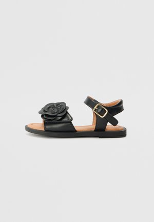 Schwarze Ledersandalen mit floralem Akzent an der Zehe, verstellbarem Knöchelriemen, flachem Sohlen und beigem Innenleben.