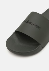 Infradito verde scuro con suola in gomma testurizzata e una cintura liscia con il nome del marchio embosso "Calvin Klein."