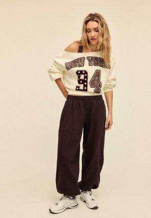 Roomkleurige sweatshirt met "NEW YORK 94" opdruk en sterren, gecombineerd met donkerbruine broek met wijde pijpen en witte sneakers met zwarte accenten.