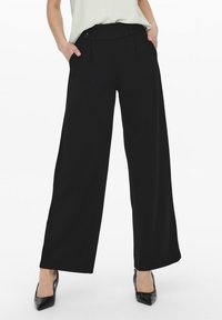 Pantalons larges noirs avec une texture lisse, présentant une taille haute, des plis devant et des poches latérales, associés à des chaussures à talons hauts noires.